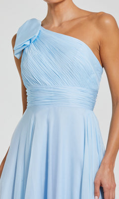 A woman wears the Mac Duggal 67476—an elegant light blue, long one-shoulder prom dress featuring pleats and a shoulder bow detail—perfect for any formal event.