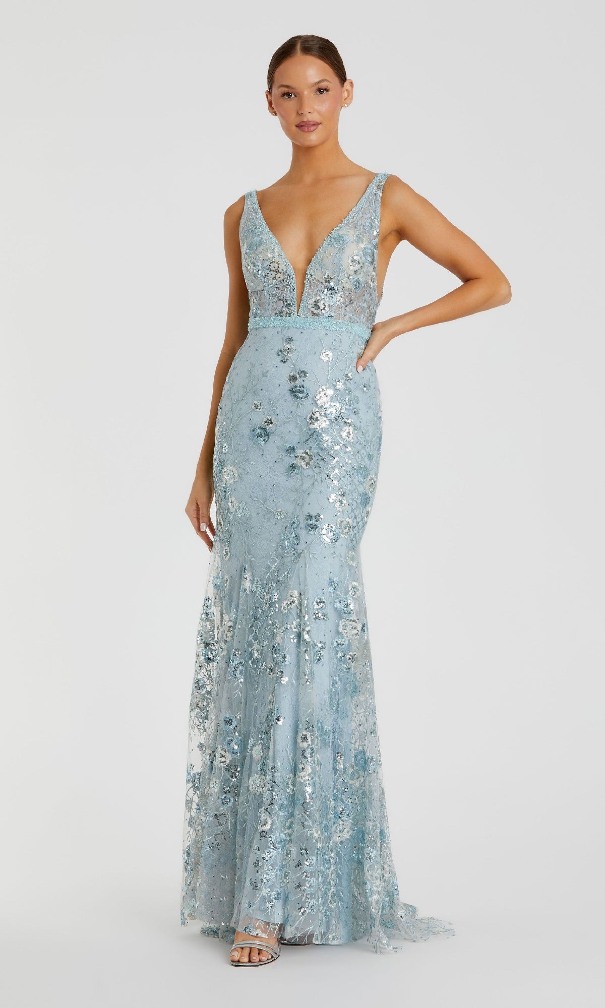 A woman stands against a plain background wearing the Mac Duggal 67481, a sleeveless long floral-print formal dress in light blue with a deep V-neckline.