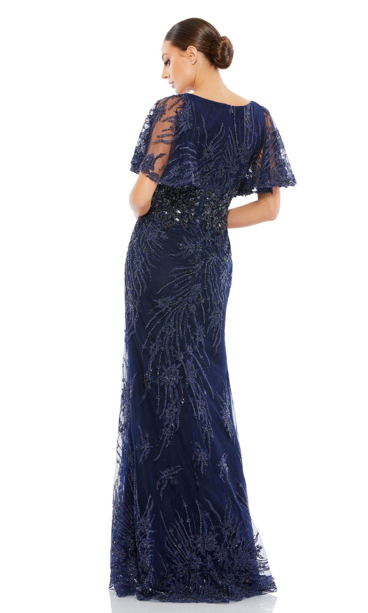 A woman stands facing away from the camera in the Long Formal Dress 67493 by Mac Duggal—a navy blue gown with a sheer beaded overlay, cape sleeves, and an empire waist.