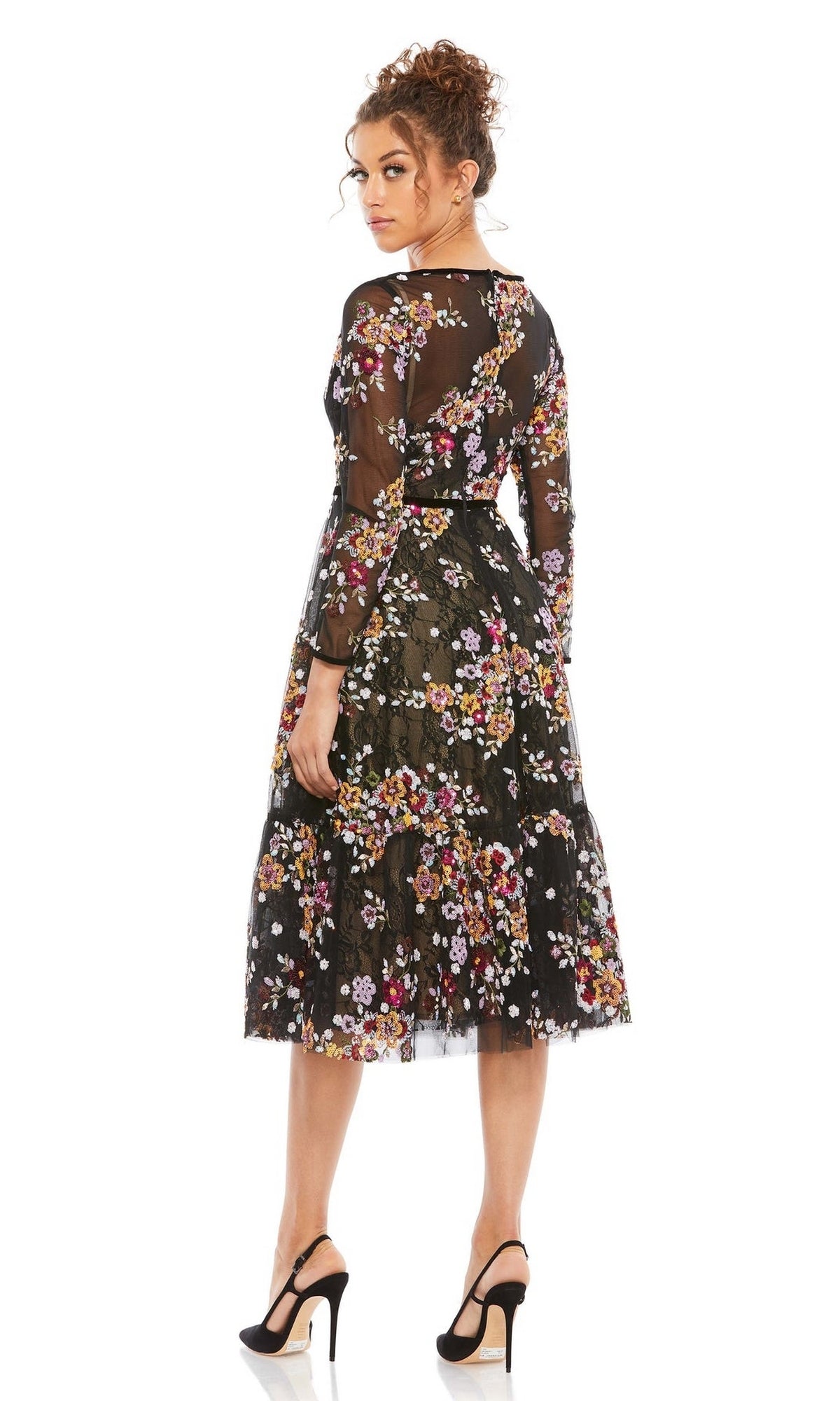 A woman in the Mac Duggal 67499 Print Midi Wedding-Guest Dress, featuring black sheer long sleeves and floral print, stands with her back to the camera, head turned, paired with black high heels.
