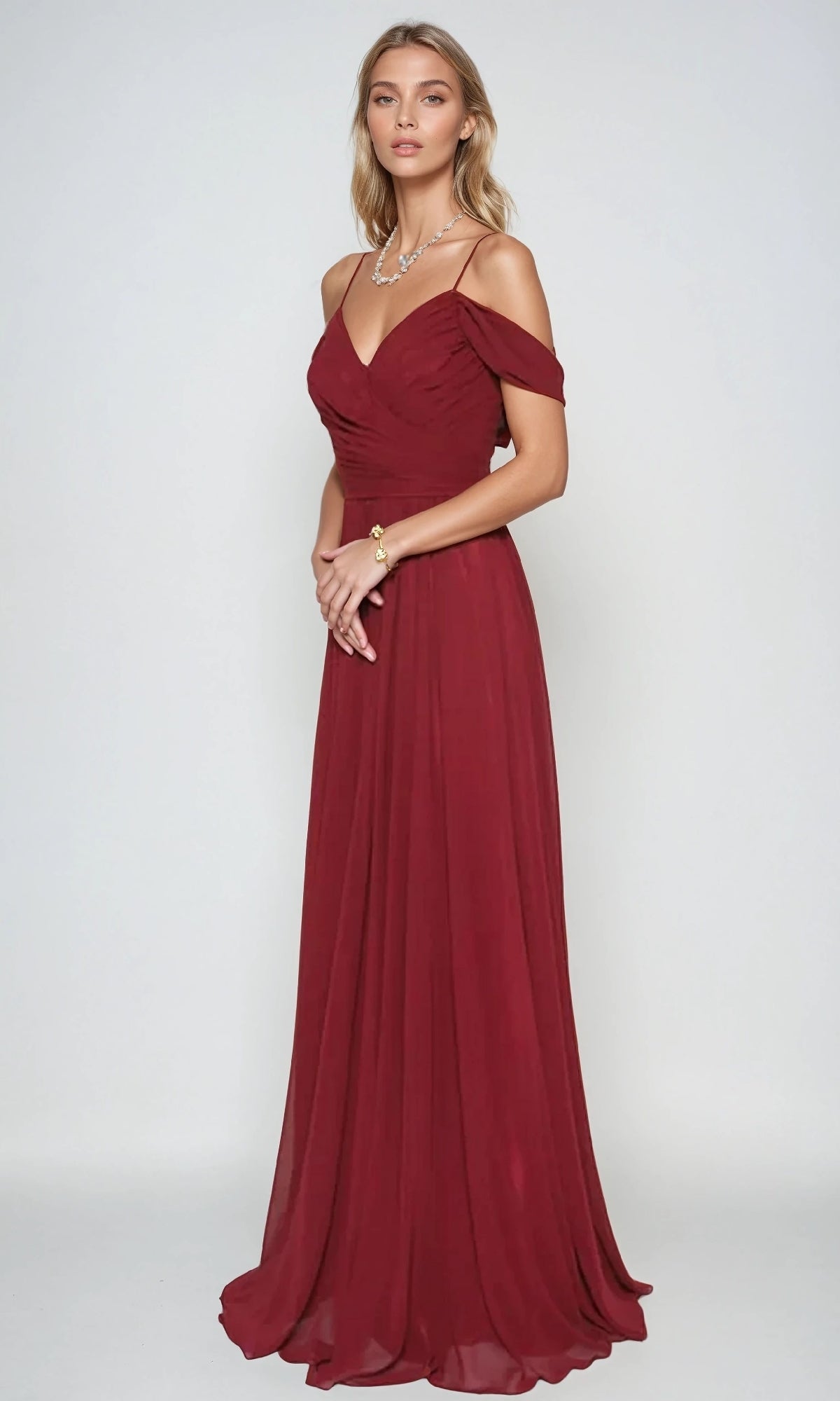 A woman wears the Juliet 676 long prom dress, featuring a floor-length, off-the-shoulder burgundy chiffon design with a fitted bodice and flowing A-line skirt, accessorized with a necklace and bracelet.