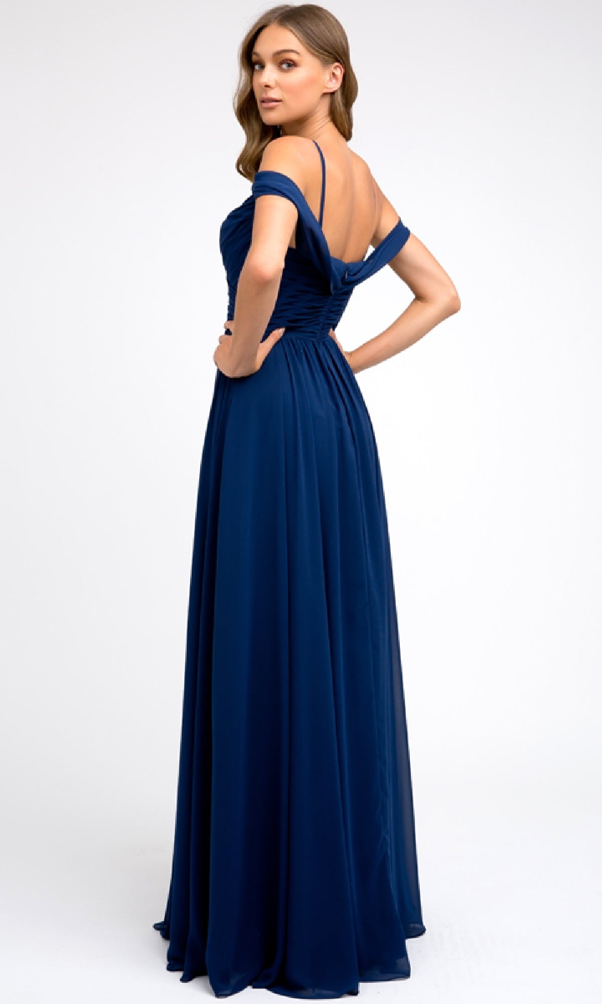 A woman models the Juliet 676 Long Prom Dress, a floor-length, off-the-shoulder navy blue gown with an elegant A-line skirt, standing with her back slightly turned against a plain white background.