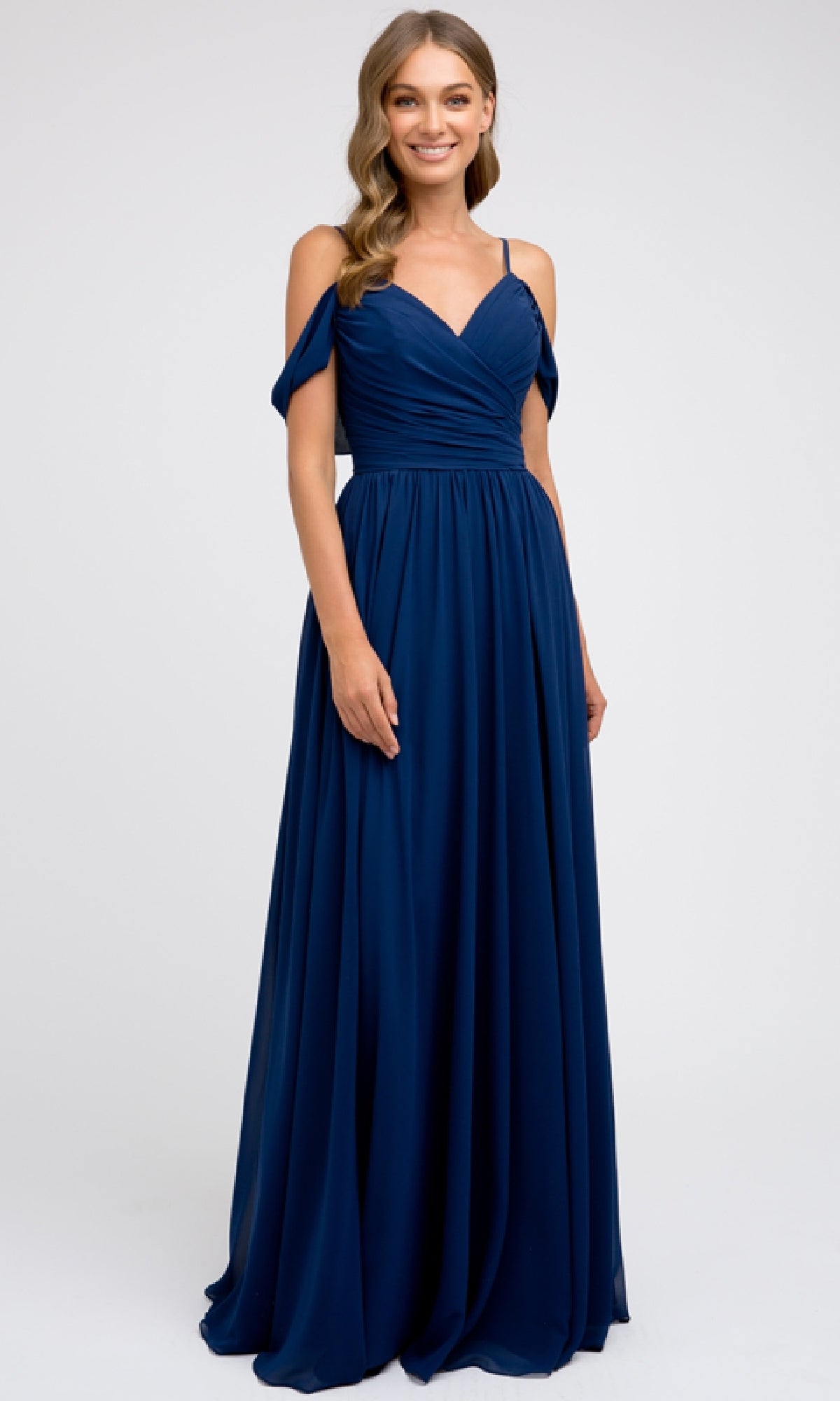 A woman wears the Long Prom Dress: Juliet 676, a floor-length dark blue chiffon gown with spaghetti straps, off-the-shoulder sleeves, and an elegant a-line skirt, standing against a plain light background.