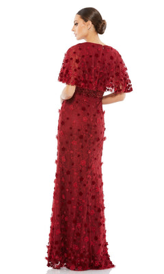 Woman poses with her back to the camera against a white background, wearing the Mac Duggal 67712 short-sleeve long formal dress, featuring textured red fabric, floral appliqués, and a cape overlay.
