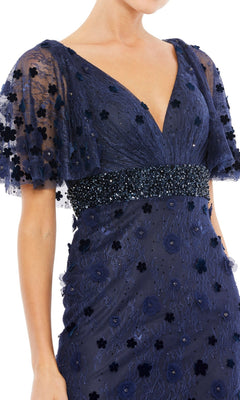 A woman wearing the Mac Duggal 67712 short-sleeve long formal dress in navy blue, featuring sheer sleeves, floral appliqué details, and a beaded waistband.