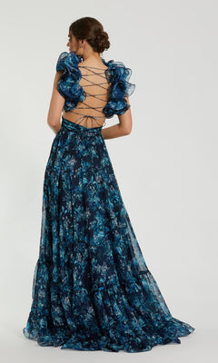 A woman stands facing away from the camera in the Mac Duggal 67803 Ruffled Long Print Formal Dress, a dark blue floral gown featuring ruffled sleeves and an open lace-up back, set against a plain background.