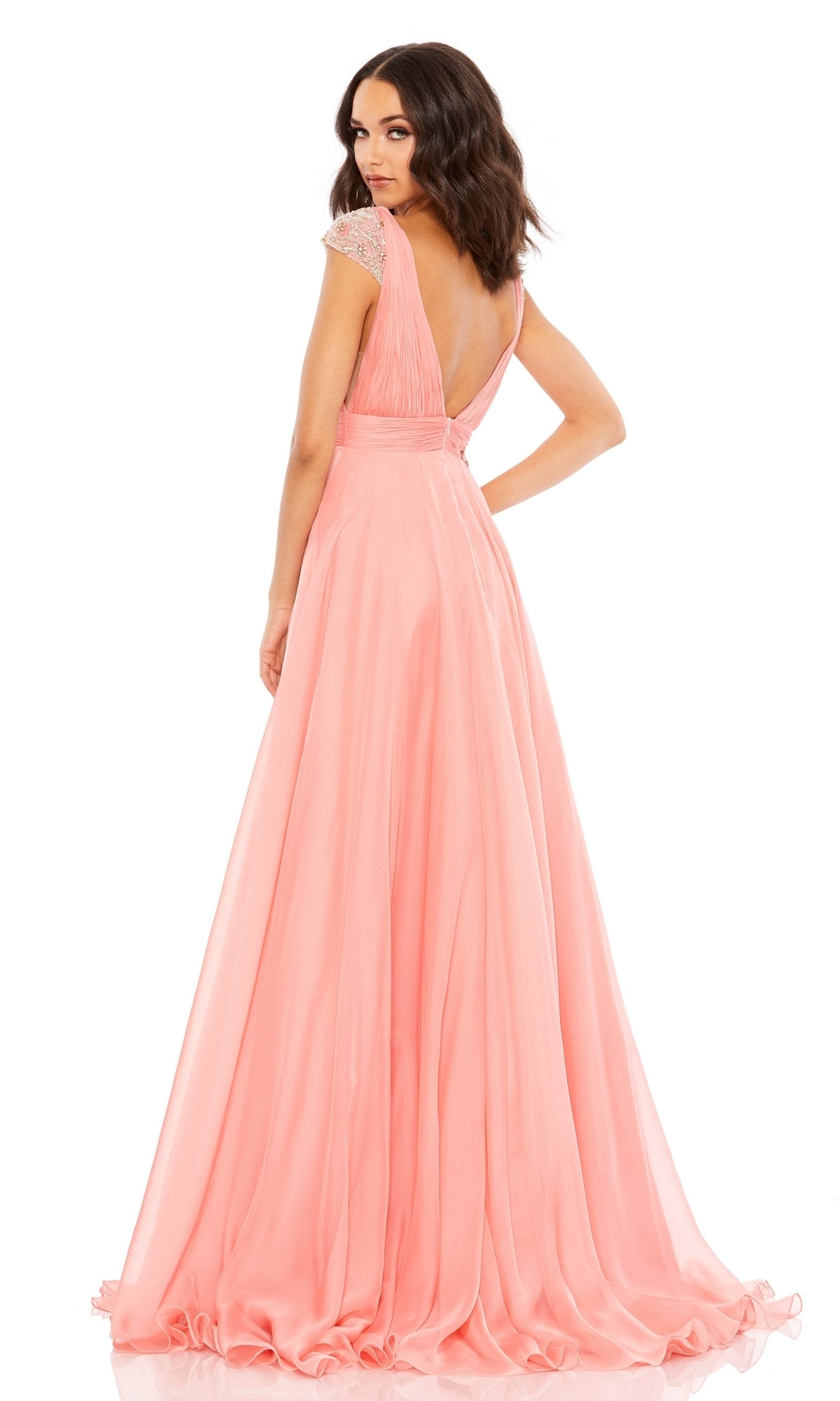 Woman in the Coral Pink Cap Sleeve Long Prom Gown 67811, featuring beaded cap sleeves and a deep V-back, stands looking over her shoulder.