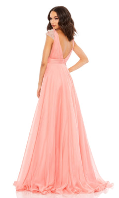 Woman in the Coral Pink Cap Sleeve Long Prom Gown 67811, featuring beaded cap sleeves and a deep V-back, stands looking over her shoulder.