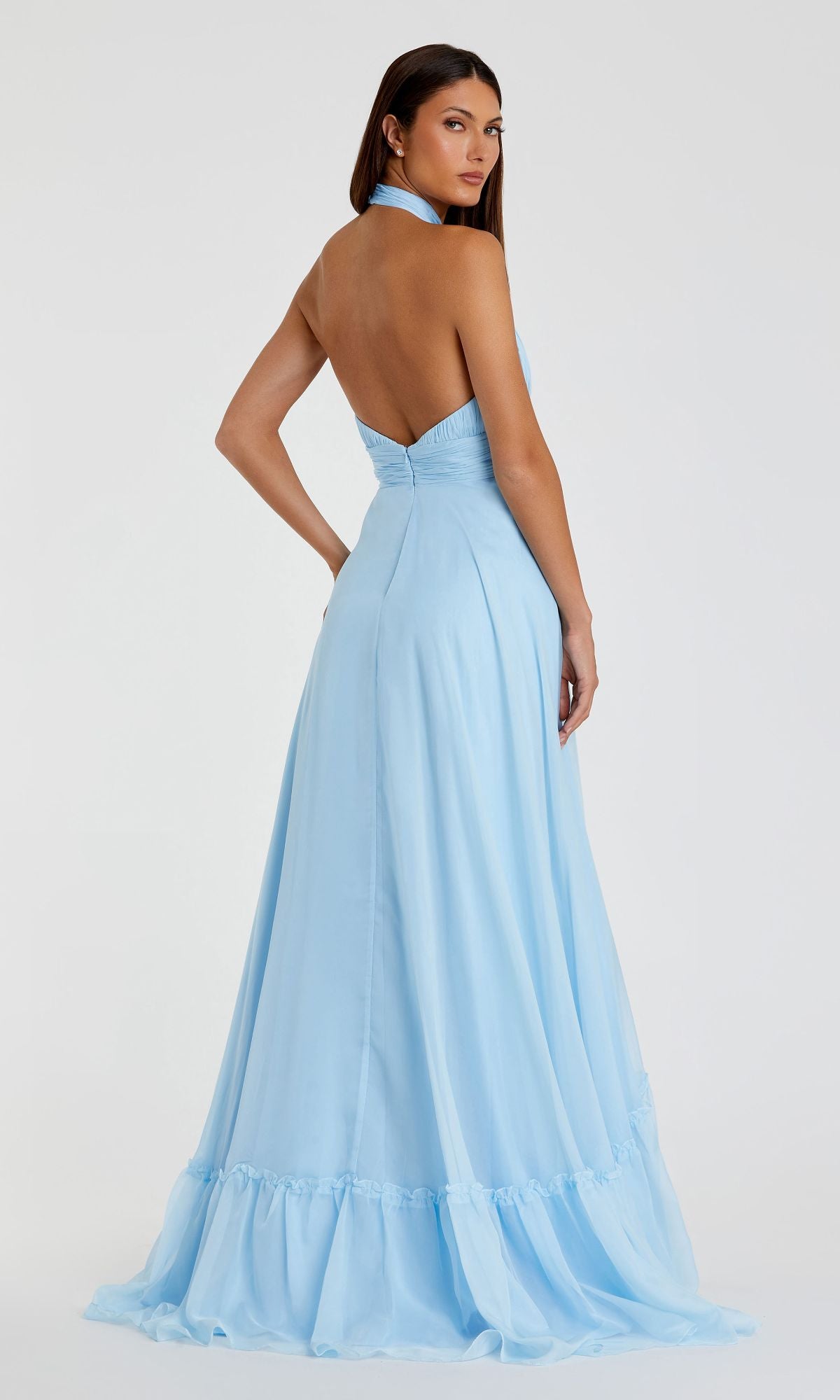 A woman wearing the Mac Duggal 67816 open-back halter formal dress in light blue stands facing away, looking over her shoulder.