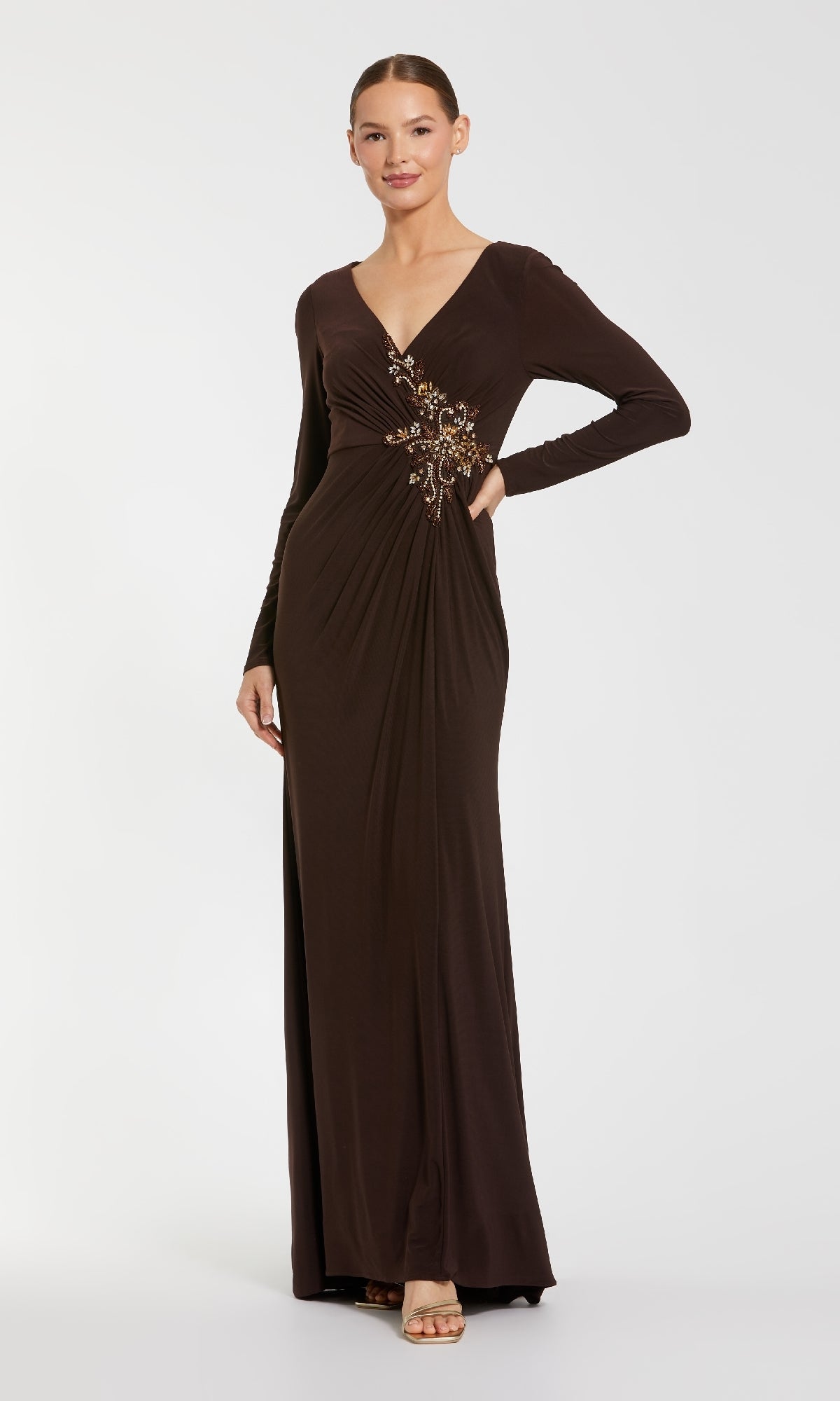 A woman poses against a plain background in the Mac Duggal 67848 Long Sleeve Long Formal Dress, a floor-length dark brown evening gown featuring a V-neck and beaded waist embellishment, with one hand on her hip.