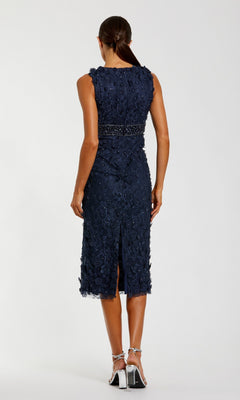A woman stands with her back to the camera in silver heels, wearing the navy blue Mac Duggal 67853 knee-length party dress featuring lace details, a beaded waistband, and a sleeveless design.