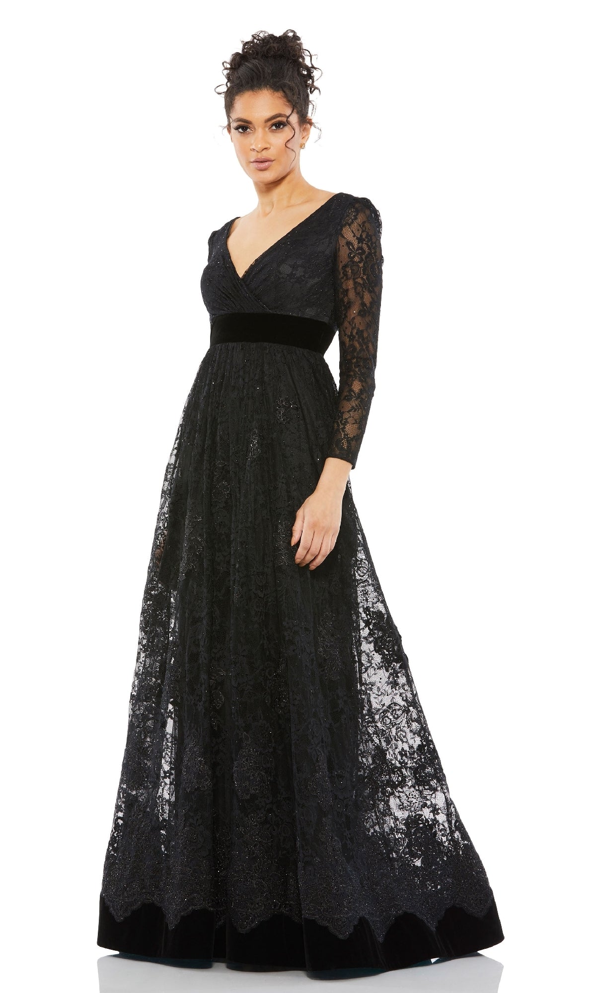 A woman stands against a white background wearing the Mac Duggal 67861 long black lace formal dress, featuring long sleeves, a V-neckline, and a velvet waistband.