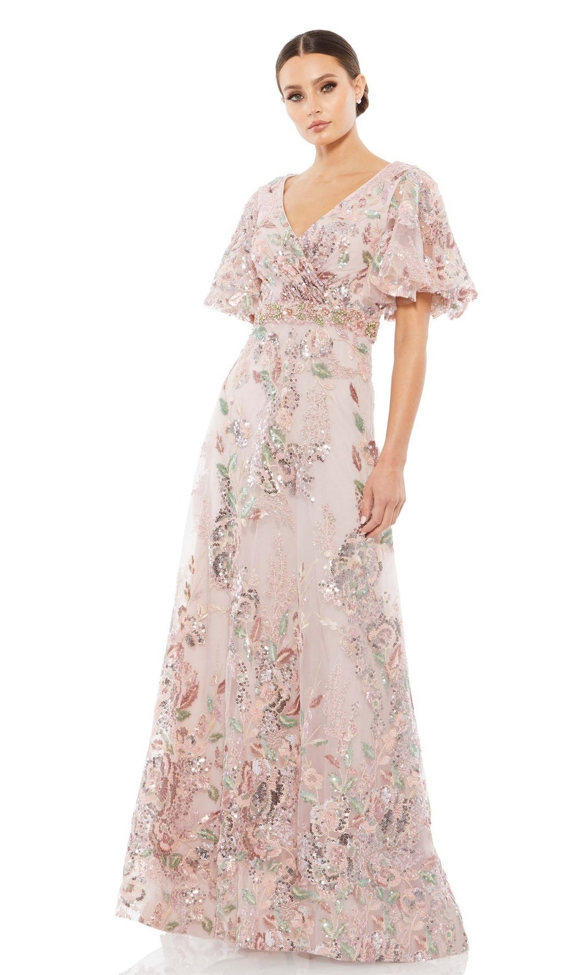 A woman wears the Mac Duggal 67880 Short Sleeve Pink Formal Dress, featuring flutter sleeves and floral sequin embellishments. This elegant gown drapes to the floor, perfect for special occasions.