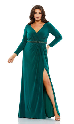 A woman poses with one hand on her hip in the Long Plus-Size Prom Dress 67899 by Mac Duggal—an emerald green gown featuring long sleeves, a v-neckline, beaded waistline, and a thigh-high slit.