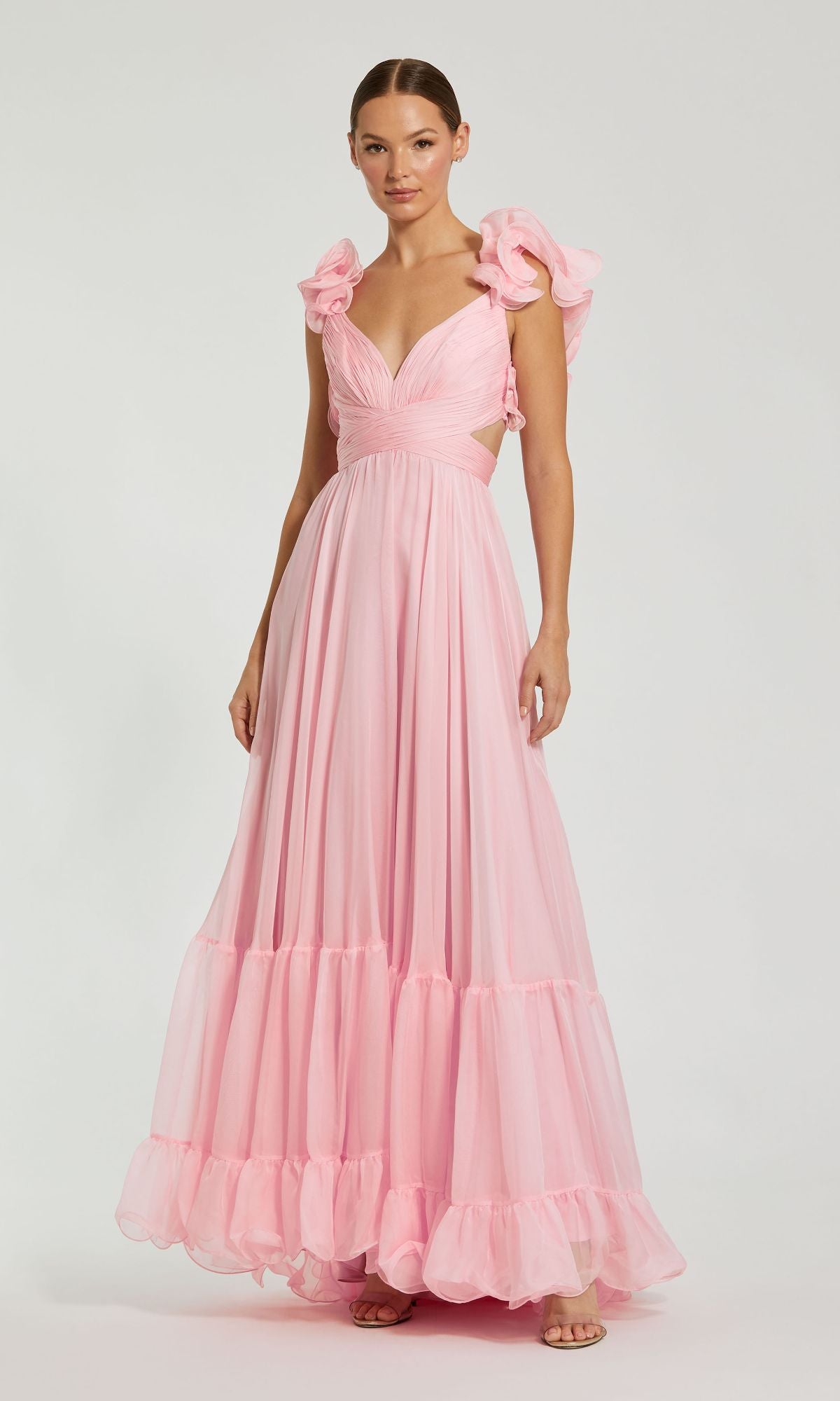A woman wears the Mac Duggal Empire-Waist Cut-Out Formal Dress 67911, a long light pink gown featuring ruffle shoulders and a deep V-neckline, standing against a plain white background.