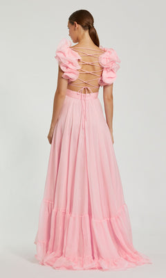 A woman stands facing away, wearing the Mac Duggal Empire-Waist Cut-Out Formal Dress 67911, a floor-length light pink gown with lace-up open back, ruffled short sleeves, and delicate detailing.