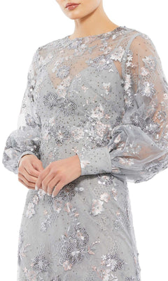 A woman stands with her hands loosely clasped, wearing the Long Formal Dress 67936 by Mac Duggal, featuring long sleeves, a floor-length silhouette, a sheer v-neckline, and intricate floral embroidery.