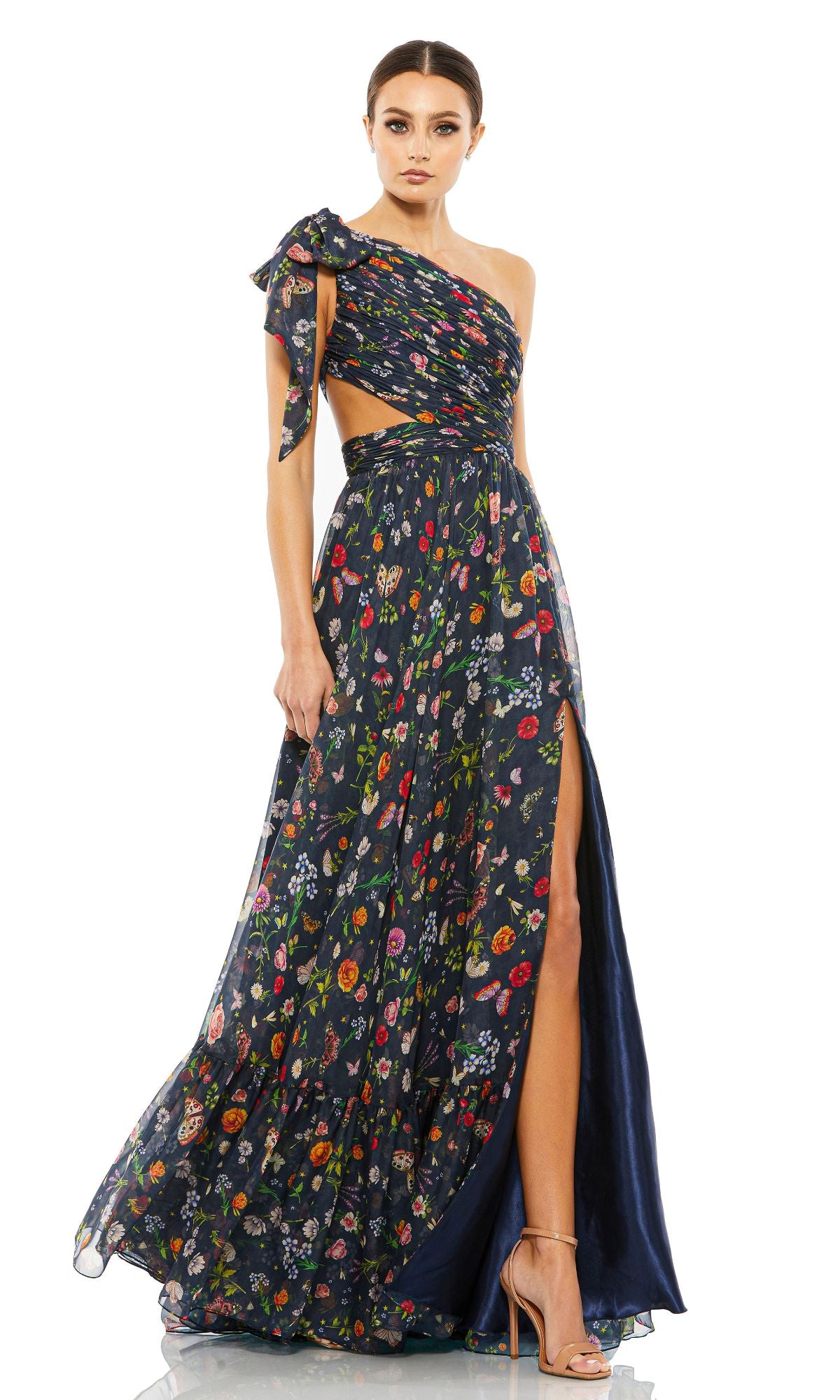 A woman wears the Long Navy Print Prom Gown: Mac Duggal 67938, a one-shoulder floral dress with a waist cutout and high slit, styled with nude high-heeled sandals.
