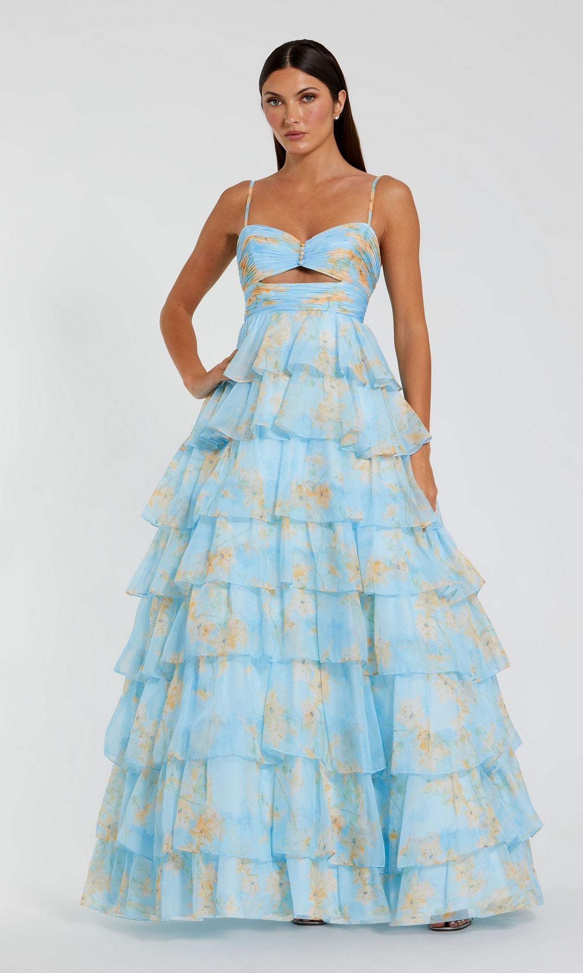 A woman stands against a plain background wearing the Mac Duggal 67966 Violet Print Prom Ball Gown, featuring ruffled tiers, thin straps, a waist cutout, and a light blue floral design.
