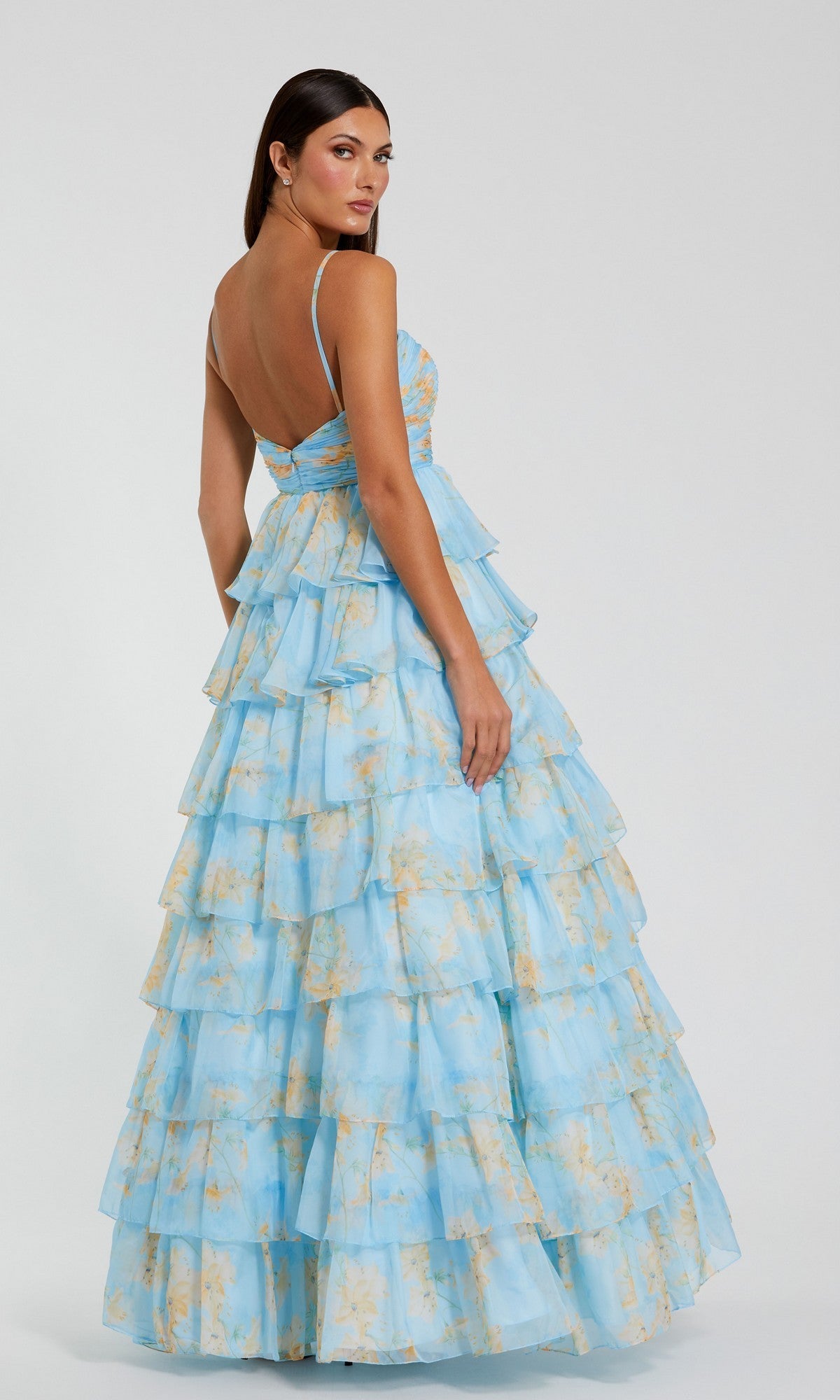 A woman faces away, wearing the Mac Duggal 67966 Violet Print Prom Ball Gown—a light blue floral ruffled tier dress with thin straps and open back—set against a simple, light background.