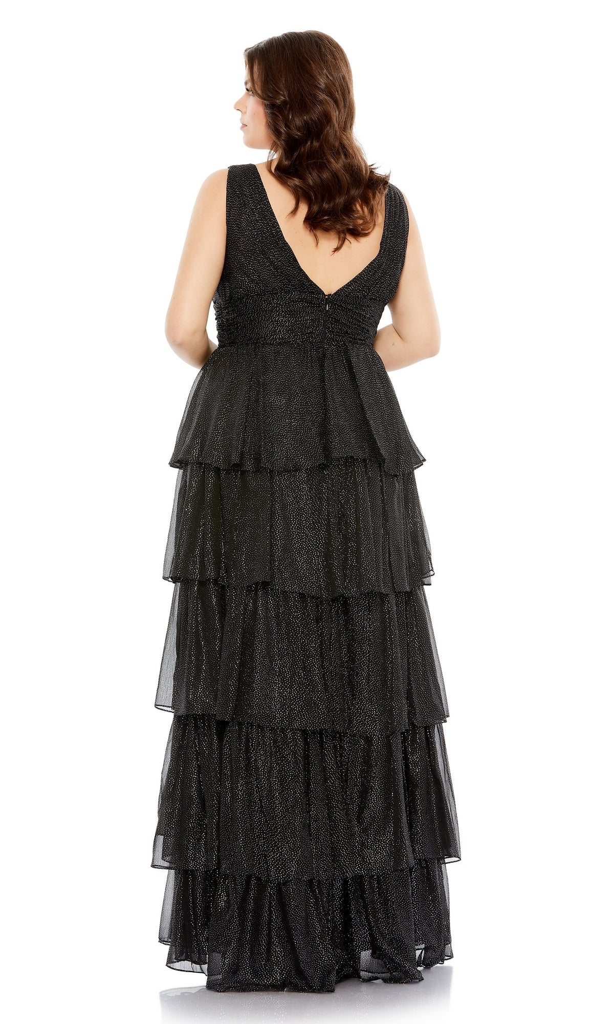 A woman with brown wavy hair is standing and facing away from the camera, wearing the Long Plus-Size Formal Dress 68131 by Mac Duggal—a sleeveless, floor-length black layered ruffle dress.