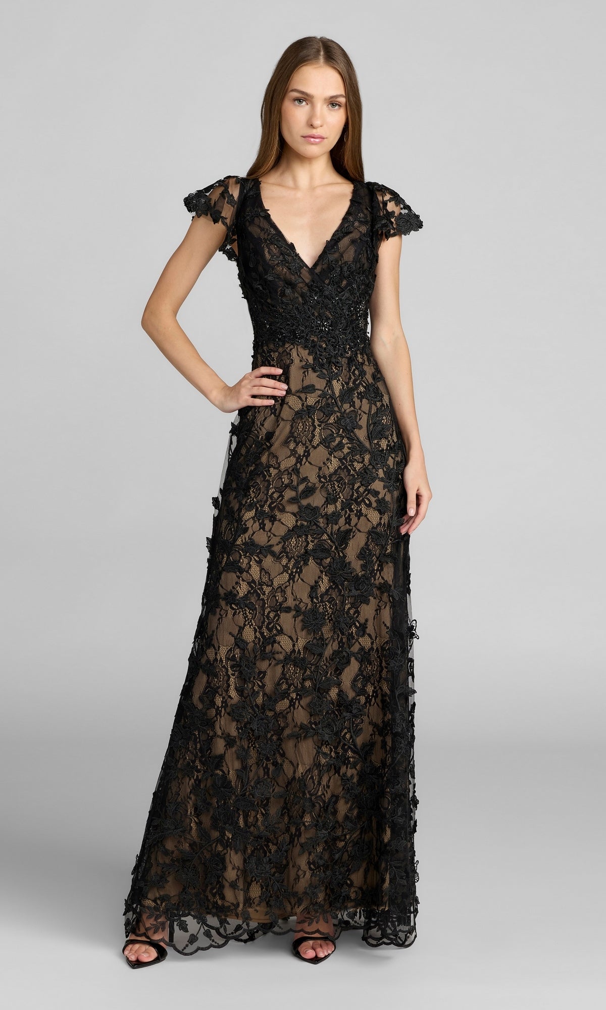 A woman poses against a plain gray background, wearing the Mac Duggal 68192 Long Beaded Formal Dress in Black Nude—a floor-length evening gown featuring short sleeves and intricate black lace with floral patterns over a nude underlay.