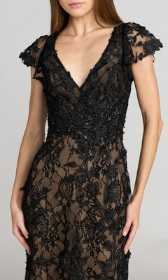 A woman wearing the Long Beaded Formal Dress: Mac Duggal 68192—a black lace gown featuring floral patterns and short sleeves—stands against a plain light background.