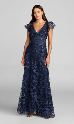Woman modeling the Mac Duggal 68192 Long Beaded Formal Dress, featuring a floor-length navy blue design with floral lace embroidery, short flutter sleeves, and a V-neckline, posed against a light gray background.