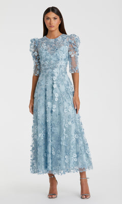 A woman wears the Mac Duggal 68193 tea-length lace formal dress in light blue, featuring puffed sleeves and sheer details, styled with silver high-heeled sandals.