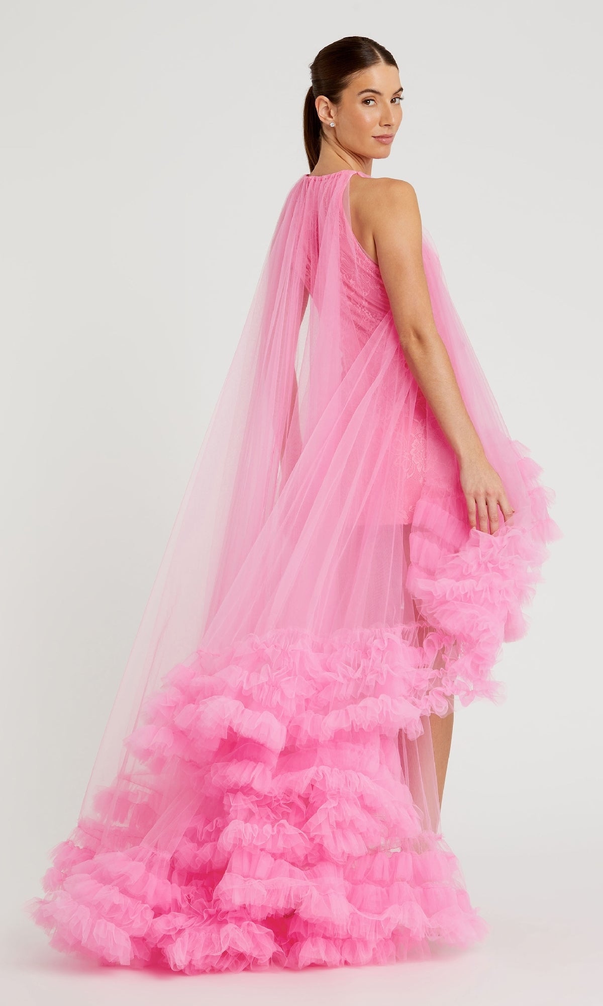 A woman poses in the Mac Duggal 68252 High-Low Trapeze Formal Dress, featuring a sleeveless design in bright pink with layered ruffles and a long tulle cape, set against a plain white background.