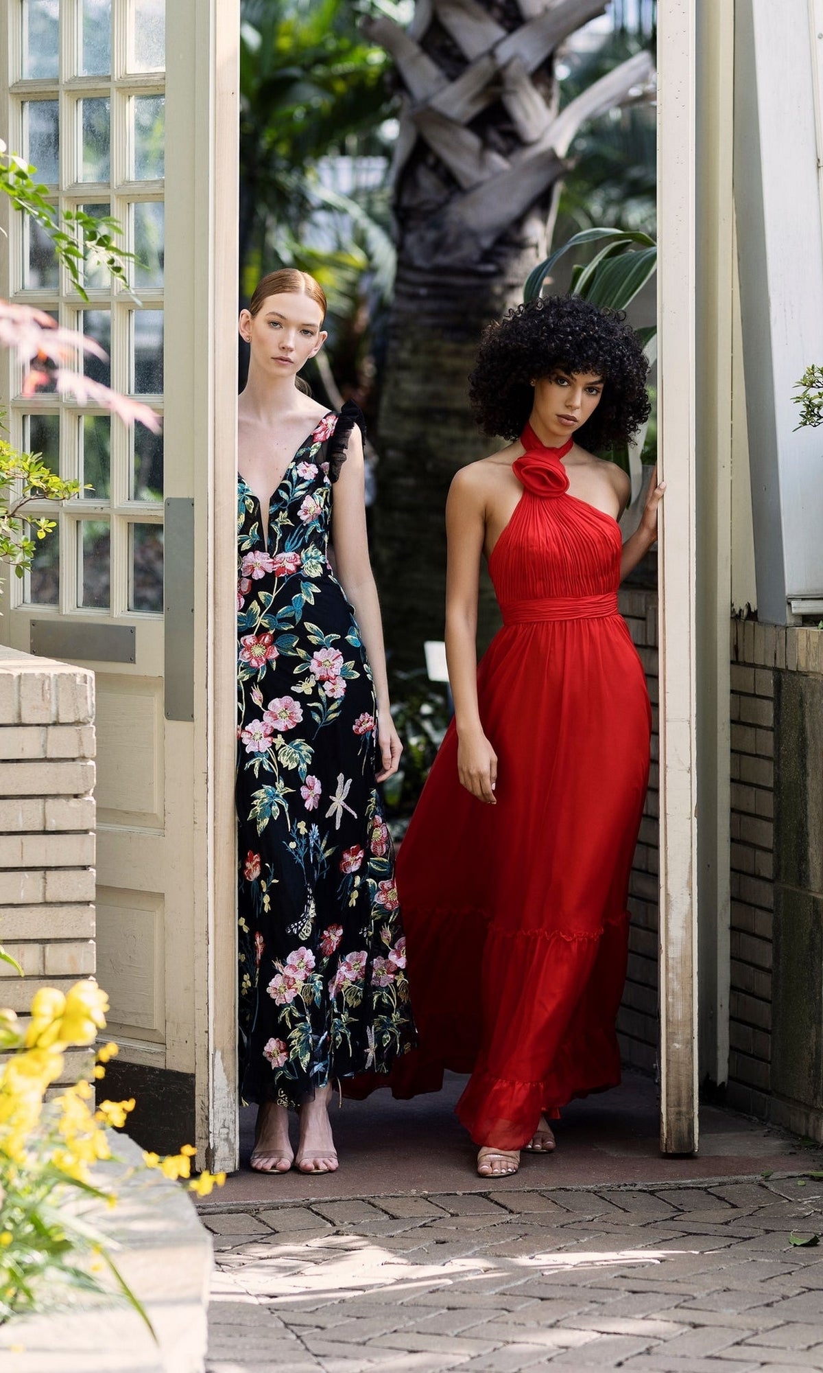 Two women stand in a doorway with greenery—one in a black floral embroidered v-neck dress, the other in the striking red Long Formal Dress 68273 by Mac Duggal, featuring a halter neckline and fitted silhouette.