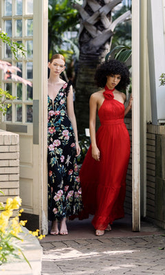 Two women stand in a doorway with greenery—one in a black floral embroidered v-neck dress, the other in the striking red Long Formal Dress 68273 by Mac Duggal, featuring a halter neckline and fitted silhouette.