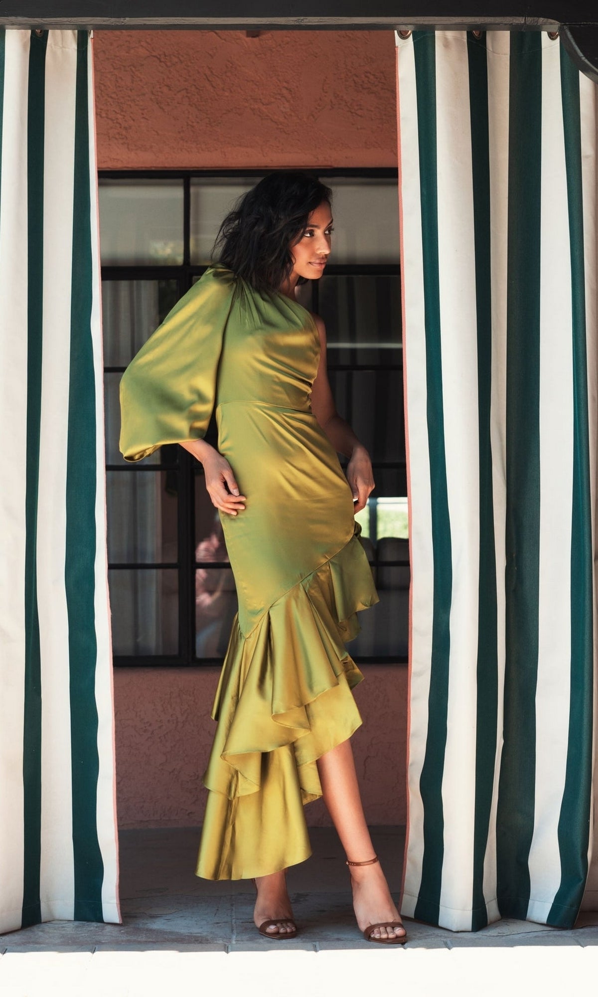 A woman wears the Mac Duggal Short Wedding Guest Dress 68330, a shiny apple green one-shoulder style with ruffled details, standing between striped curtains and looking to the side with one hand on her hip.