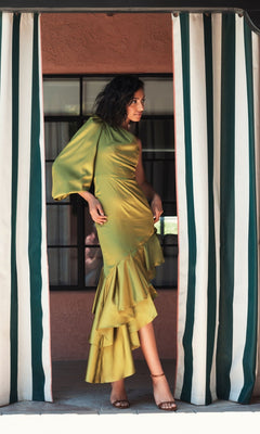 A woman wears the Mac Duggal Short Wedding Guest Dress 68330, a shiny apple green one-shoulder style with ruffled details, standing between striped curtains and looking to the side with one hand on her hip.