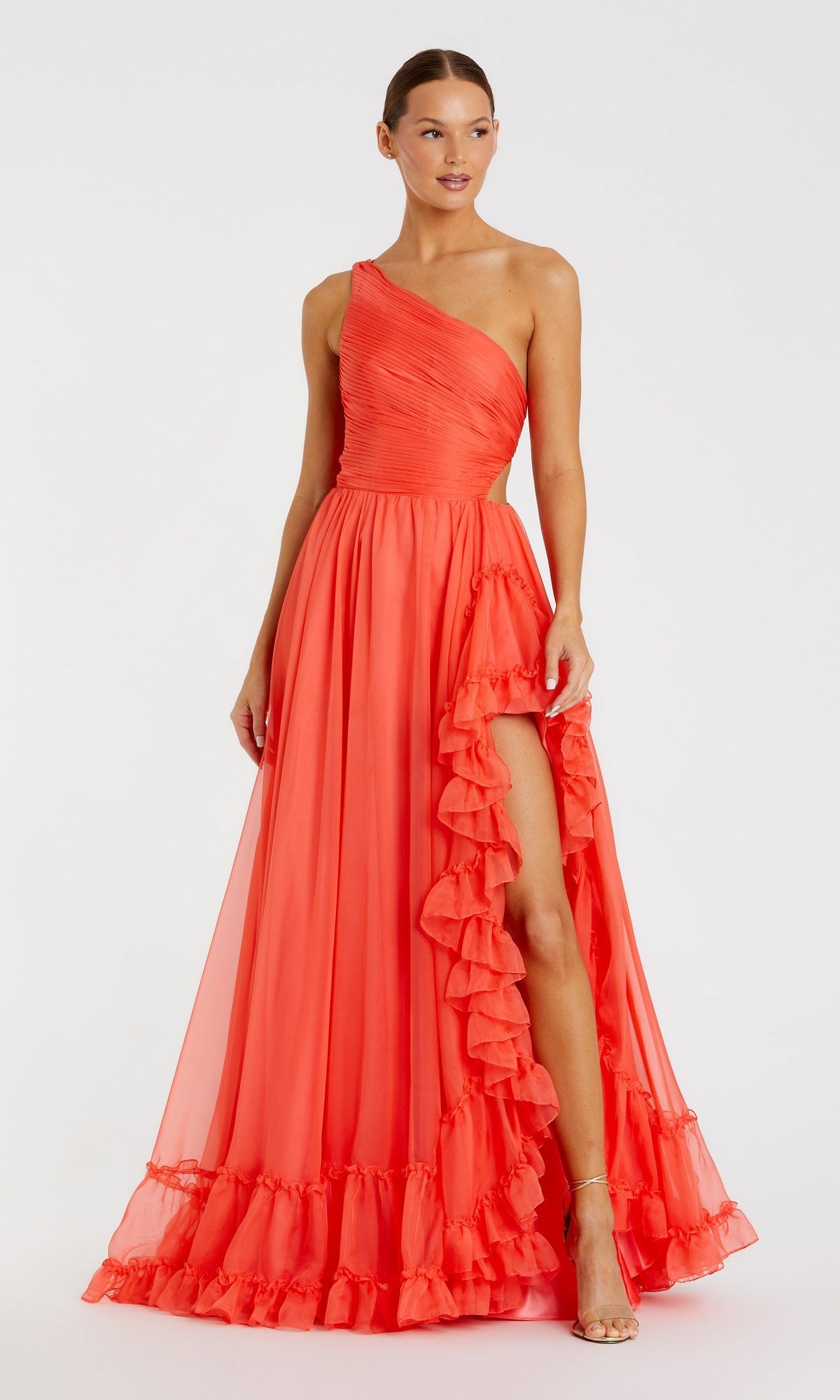 A woman wears the Mac Duggal 68476 long formal dress in tangerine, featuring a one-shoulder design with ruffled trim and a high slit, standing against a plain white background.