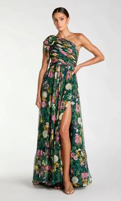 A woman poses against a plain background in the Mac Duggal 68553 long formal dress, a deep green multi floral chiffon one-shoulder evening gown with a side slit and bow detail on the shoulder.