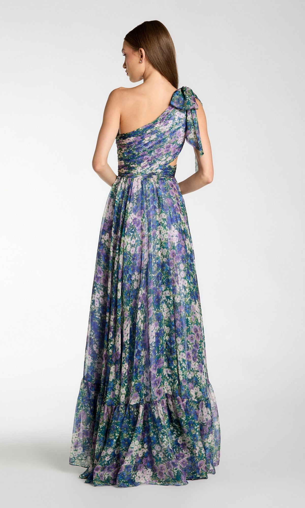 A woman faces away, wearing the Mac Duggal 68553 One-Shoulder Floral Formal Gown with a bow on the strap. The long A-line dress features a flowy layered skirt in blue, purple, and green shades.