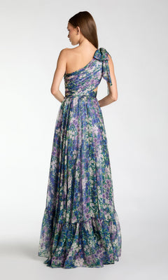 A woman faces away, wearing the Mac Duggal 68553 One-Shoulder Floral Formal Gown with a bow on the strap. The long A-line dress features a flowy layered skirt in blue, purple, and green shades.