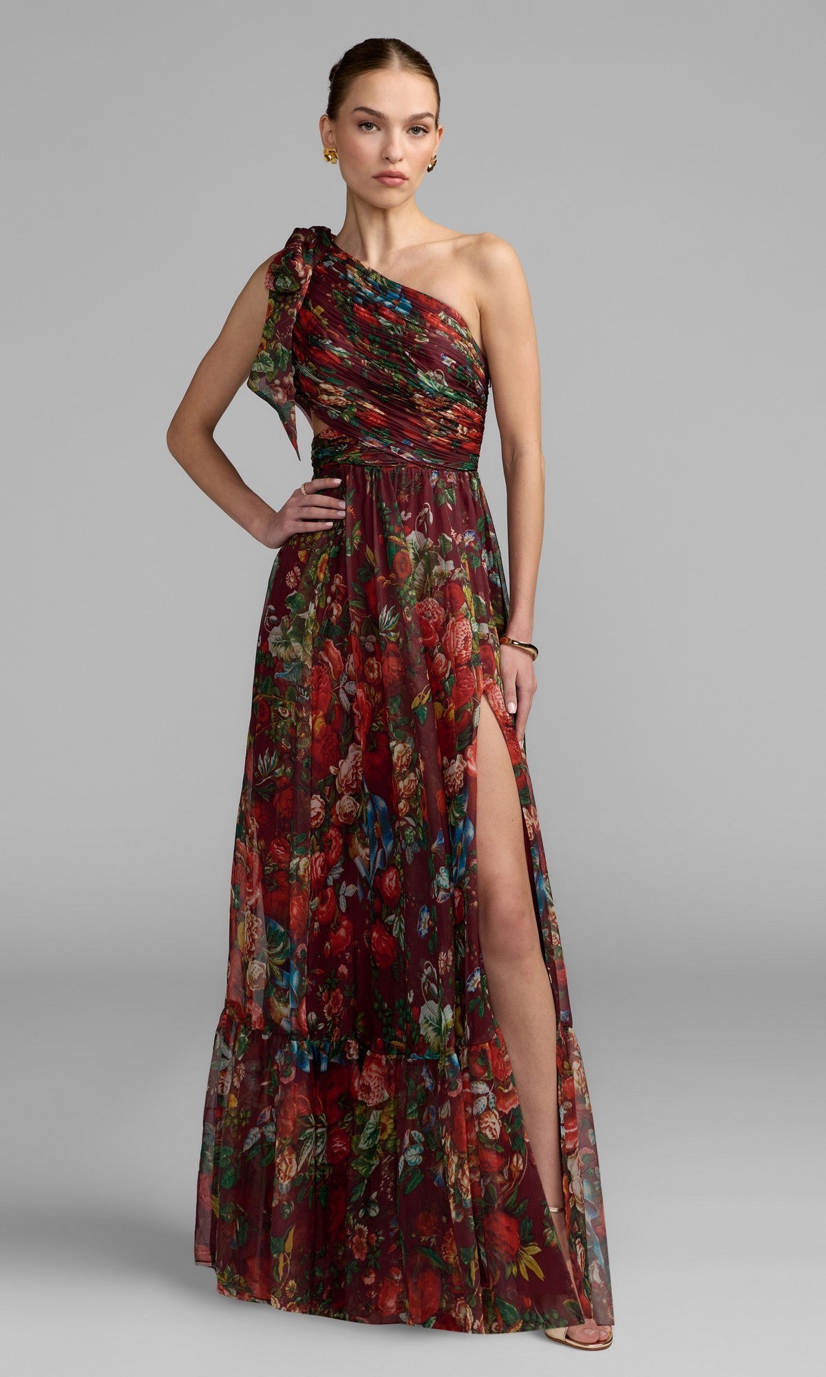 A woman models the Mac Duggal 68553 One-Shoulder Floral Formal Gown, featuring a red and green pattern and thigh-high slit, against a plain gray background.