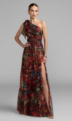 A woman models the Mac Duggal 68553 One-Shoulder Floral Formal Gown, featuring a red and green pattern and thigh-high slit, against a plain gray background.