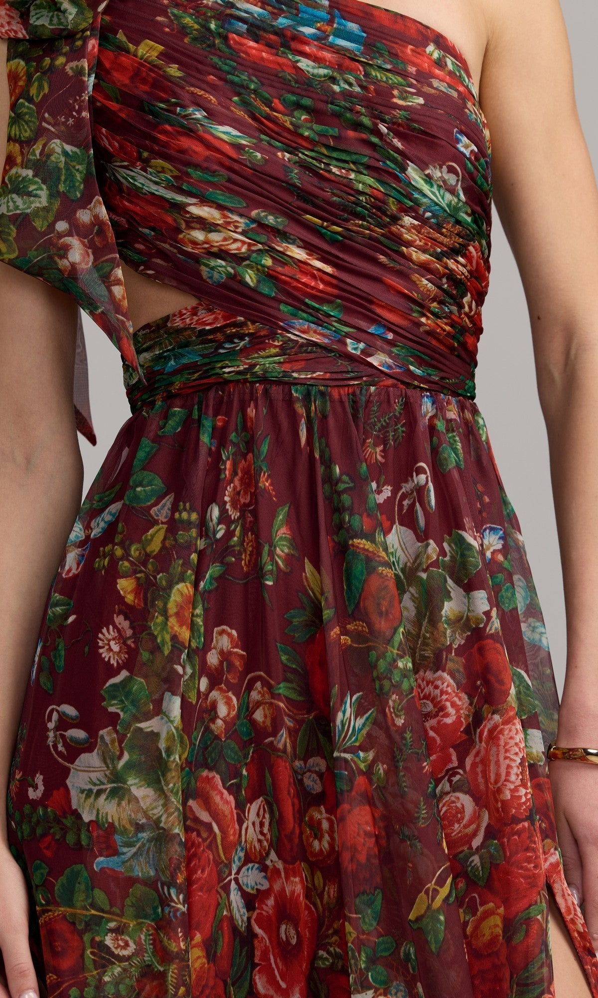 A close-up of a person wearing the One-Shoulder Floral Formal Gown: Mac Duggal 68553, featuring a maroon hue, colorful floral print, and sheer fabric overlay.