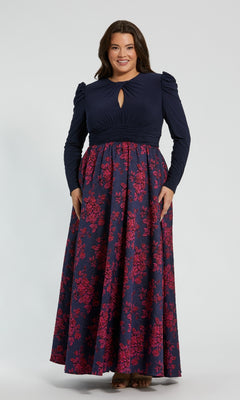 A woman models the Mac Duggal 68556 plus-size long blue formal dress, featuring long sleeves, a keyhole neckline, and a dark floral skirt, standing before a plain light background.