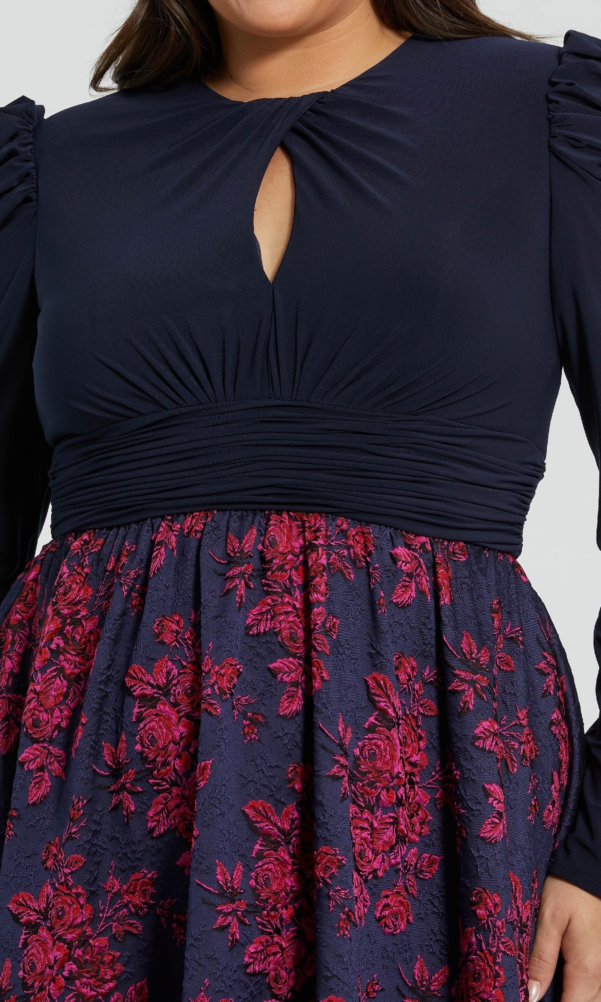 A woman wearing the Mac Duggal 68556 plus-size long blue formal dress, featuring long sleeves, a ruched waistband, a keyhole neckline, and a skirt with pink floral embroidery.