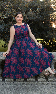 A woman wears the Plus-Size Knee-Length Print Party Dress 68557, featuring blue and pink patterns, while sitting on a stone railing outdoors with green foliage in the background.