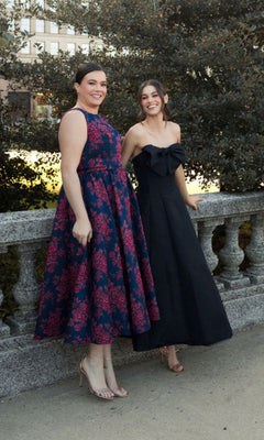 Two women pose outside by a stone railing. One smiles in the Plus-Size Knee-Length Print Party Dress 68557, while the other wears a strapless black gown; both are beaming at the camera.