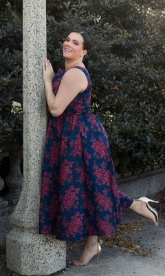 A person poses smiling by a stone lamppost in the Plus-Size Knee-Length Print Party Dress 68557, featuring red floral patterns. They wear gold high heels with one leg bent. Lush greenery appears in the background.