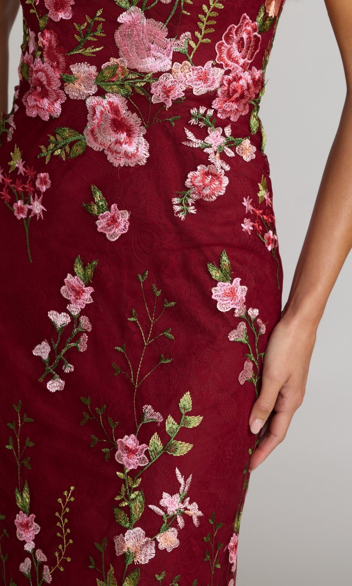 A person is shown from torso to thigh wearing the Mac Duggal 68606 long, dark red floral-print formal dress with pink and green embroidery, one hand resting at their side.