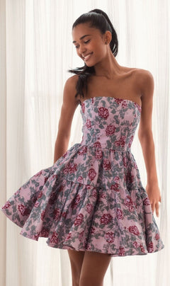 A woman smiles and looks down while wearing the Short Pink Floral Dress 68624 by Mac Duggal, featuring a strapless design with pink, grey, and red floral patterns, standing in front of sheer curtains.
