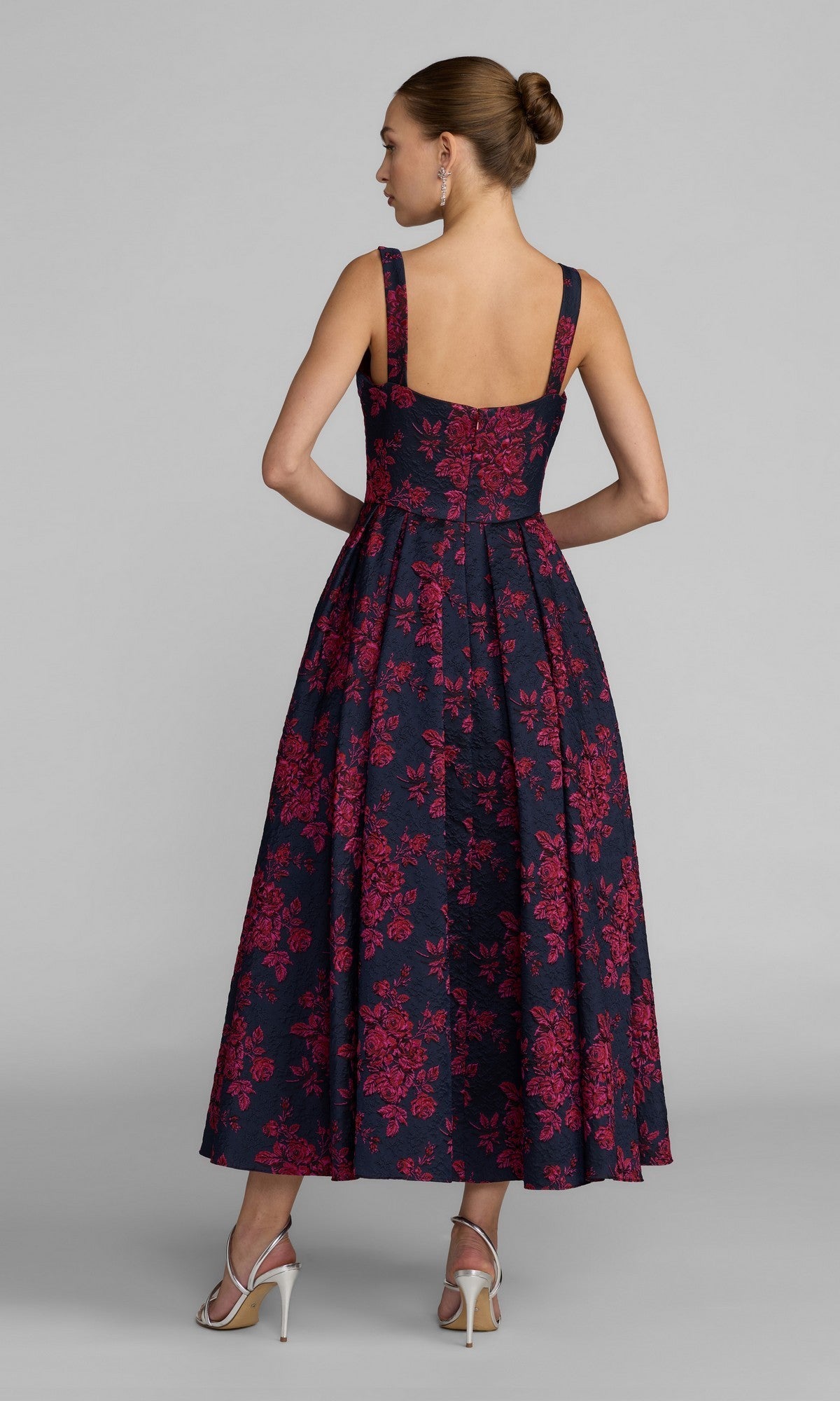 A woman, her hair styled in a bun, is facing away from the camera and wearing the Mac Duggal 68625 Tea-Length Party Dress—a sleeveless navy dress with red floral patterns—paired with white high-heeled sandals against a plain gray background.