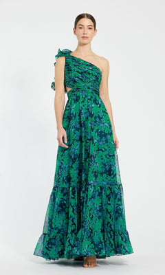 A woman stands against a white background wearing the Mac Duggal 68650 One-Shoulder Print Formal Dress, a long, sleeveless gown in green and blue floral print with ruffled detail.